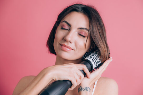 Women on pink background hold round brush hair dryer to style hair